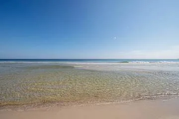 Una playa de arena se extiende junto a aguas poco profundas bajo un cielo despejado.