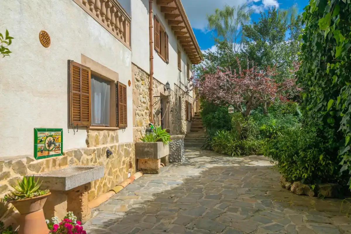 A stone-paved courtyard runs alongside a white building with wooden shutters and garden plants.