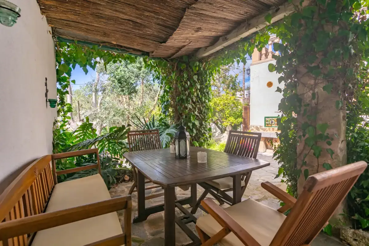 A shaded outdoor dining area with a wooden table and seating beneath a vine-covered pergola.