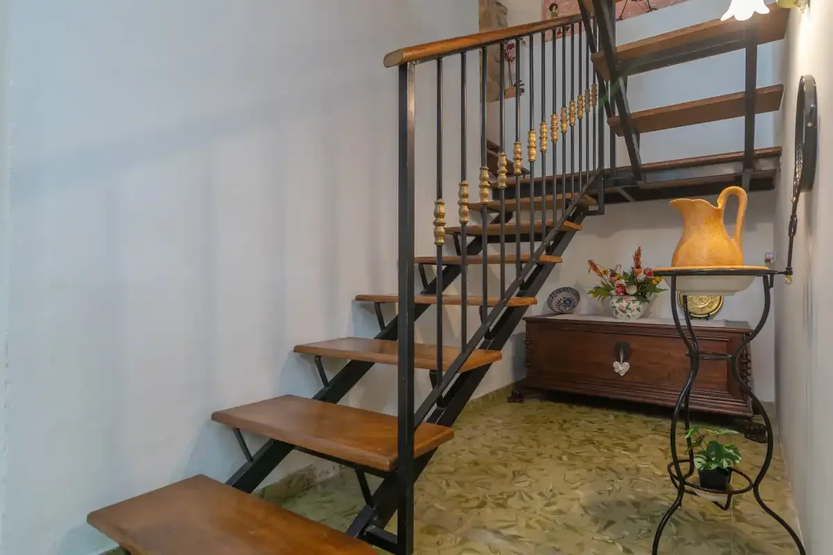 An indoor staircase with dark railings rises beside a wooden chest and decorative pitcher.