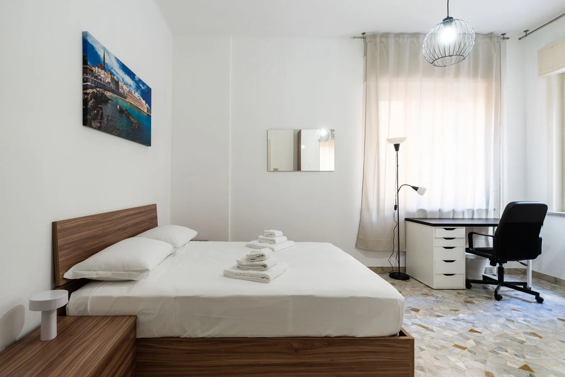 A bedroom with a bed, bedside table, and desk beside a bright window.