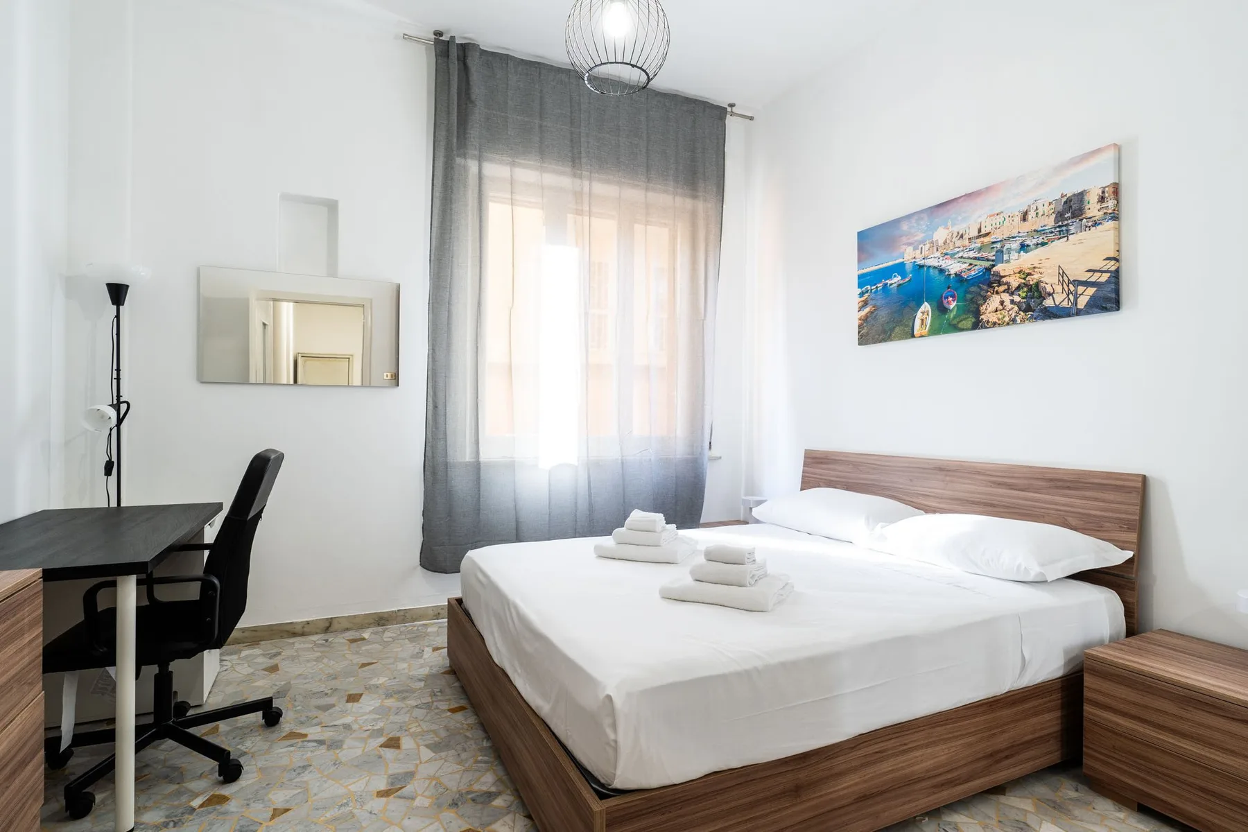A bedroom with a double bed, a desk, and a large curtained window.