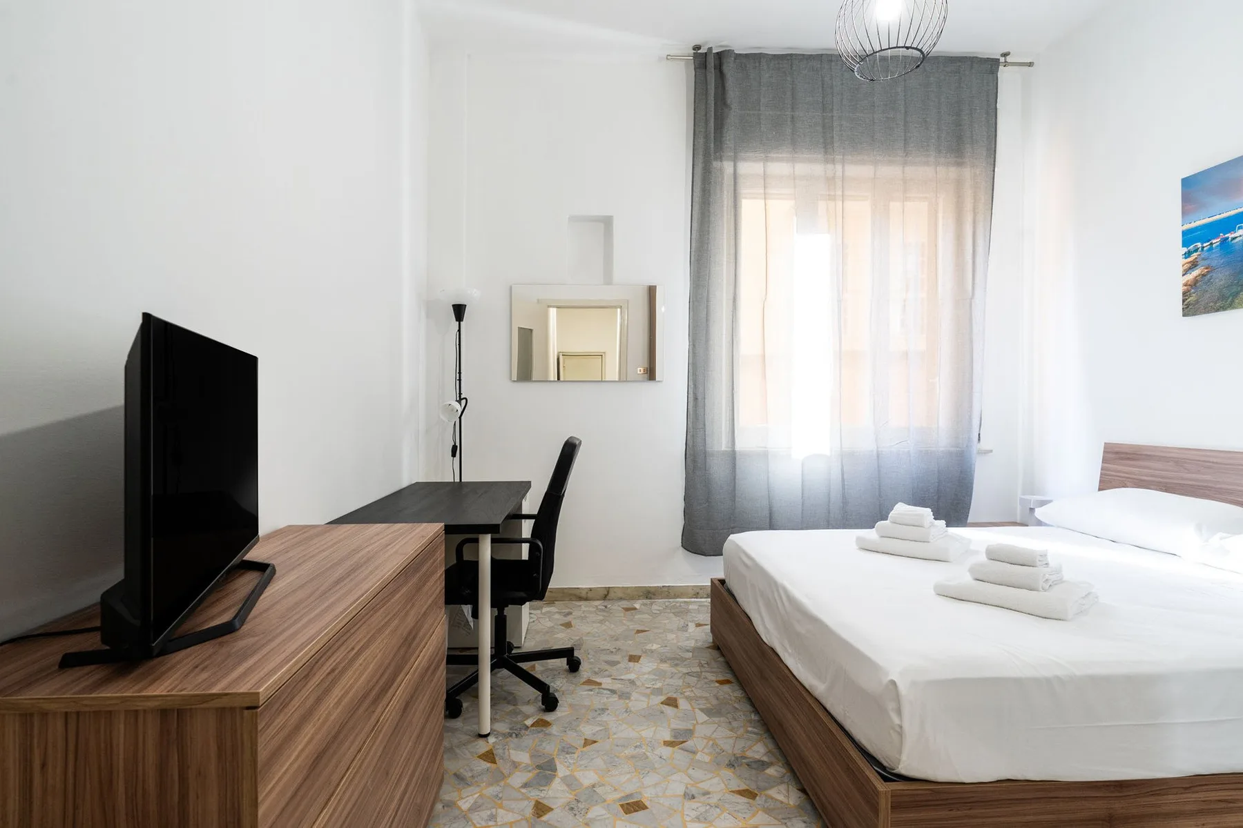 A bright bedroom with a bed, desk, and television beside a window.