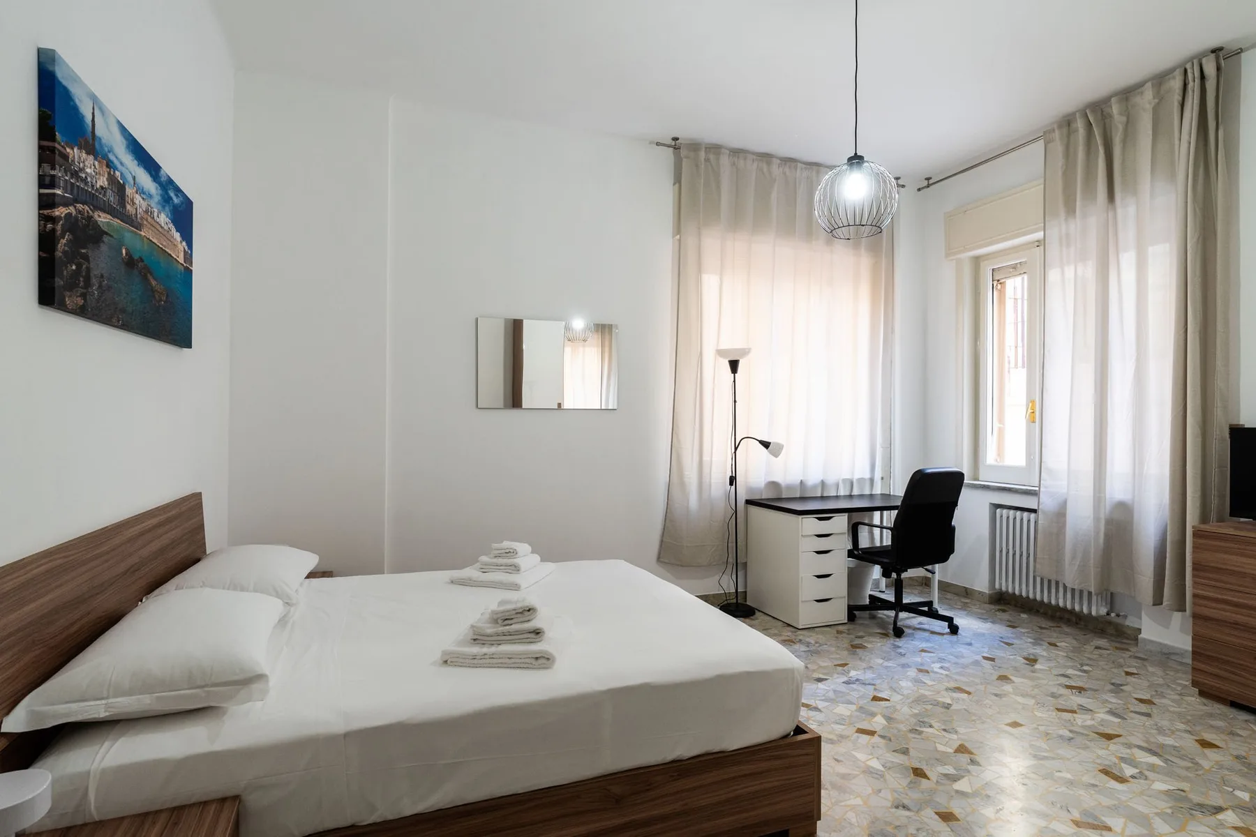 A bedroom with a bed, desk, and tall curtained windows.