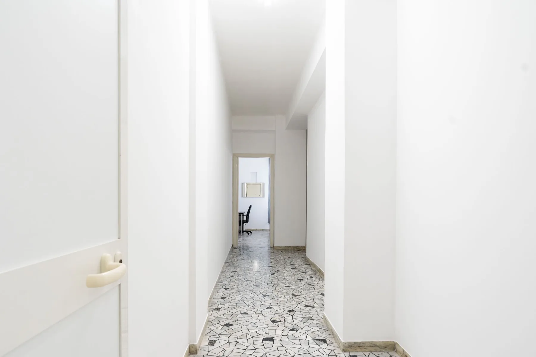 A narrow hallway with a patterned tile floor leads to a bright room.