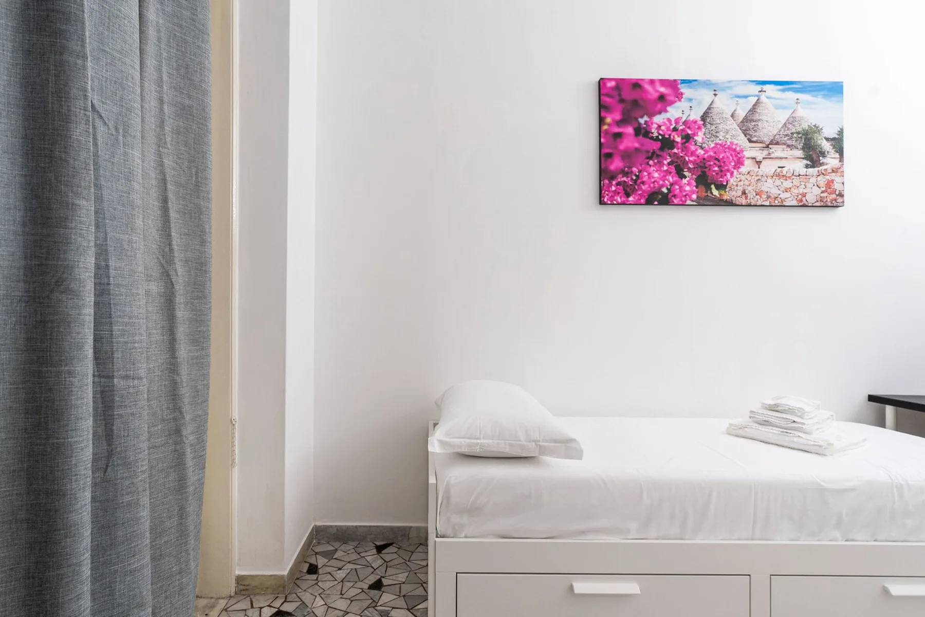 A white bed sits against a plain wall beside a gray curtain.