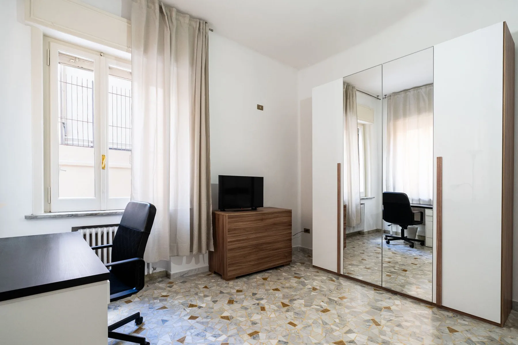 A bright room with a desk, chair, and mirrored wardrobe.