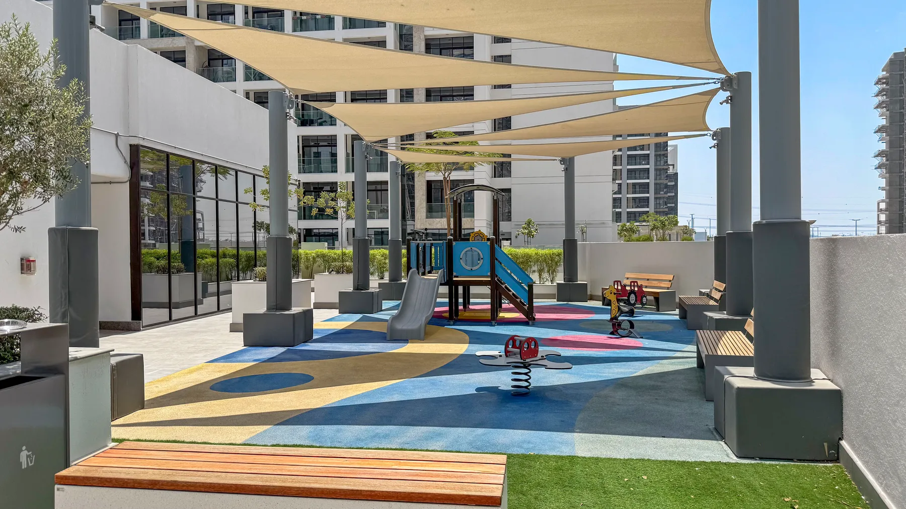 An outdoor playground with colorful rubber flooring and shade sails between buildings.