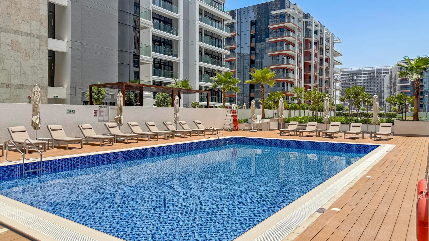 An outdoor swimming pool sits beside a wooden deck and modern apartment buildings.