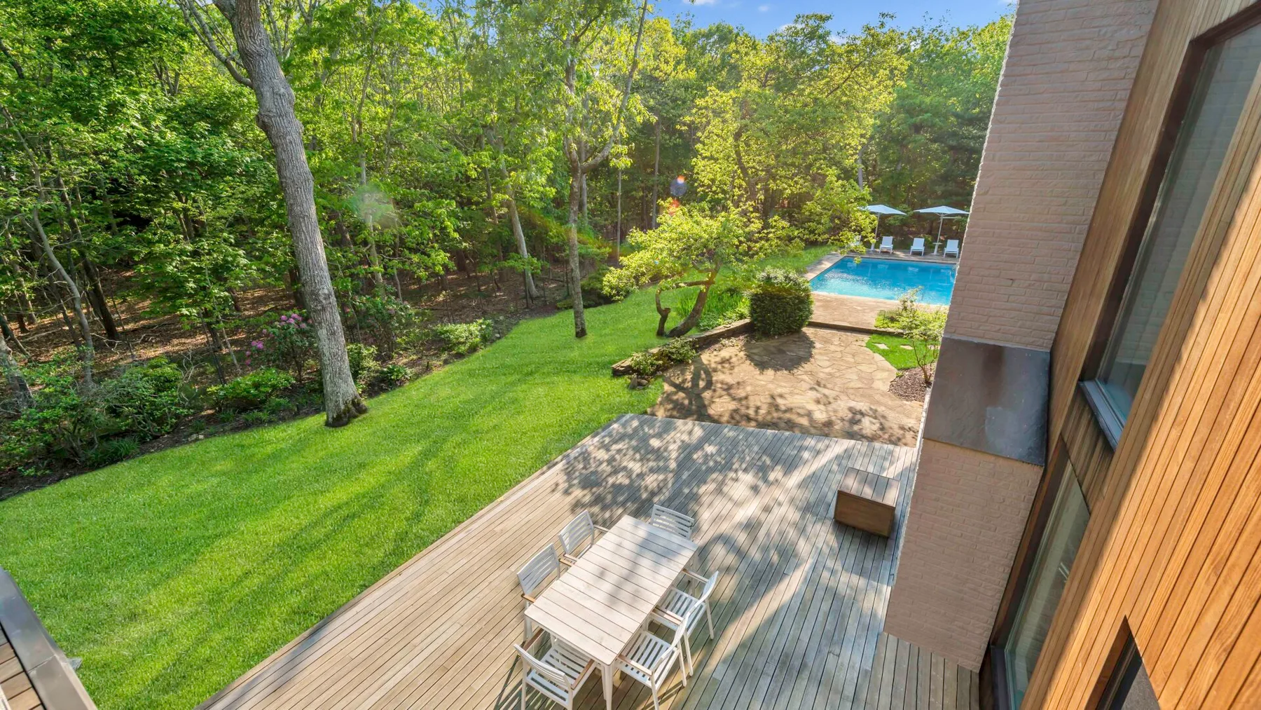 Un patio arbolado tiene césped, una terraza de madera con asientos y una piscina.