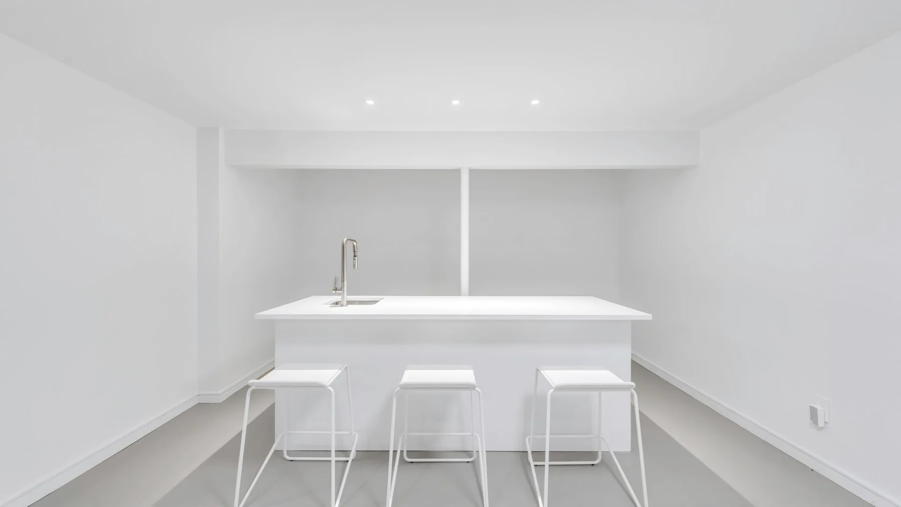 Una encimera blanca y luminosa da frente a tres taburetes en una estancia minimalista.