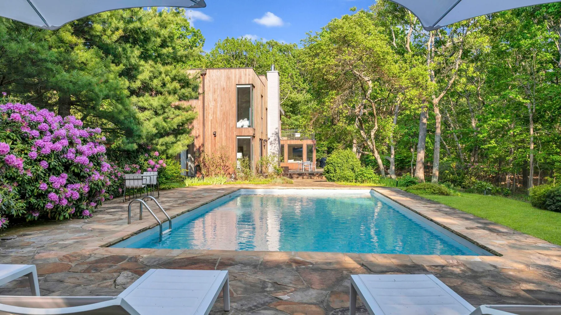 Una piscina al aire libre está rodeada de árboles y arbustos con flores junto a un edificio de madera.