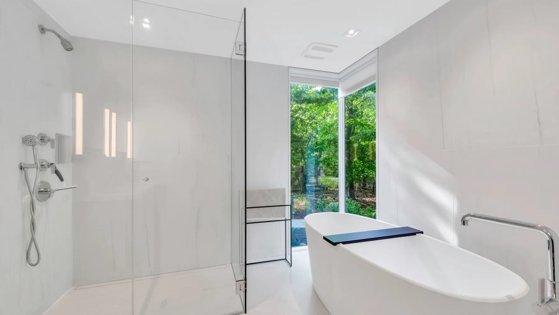 Un baño luminoso tiene una bañera junto a una ducha con mampara de vidrio y ventanas con vegetación.