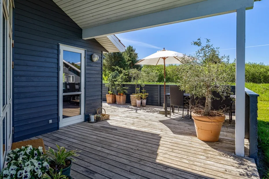 A covered wooden deck with outdoor seating, potted plants, and a sun umbrella.