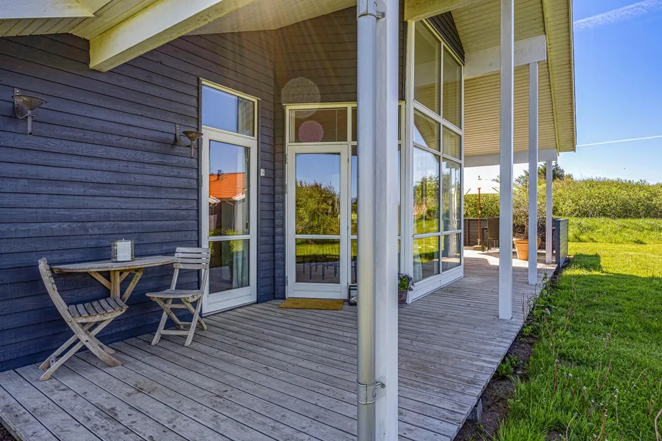 A covered wooden deck beside a house has seating and glass doors.