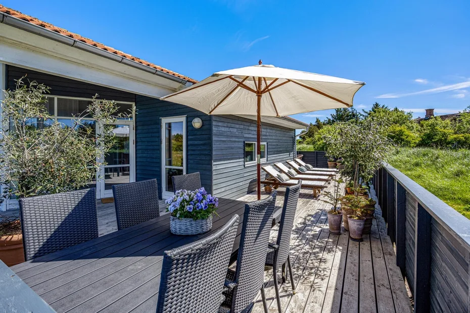 A wooden deck beside a blue house has outdoor seating and a large umbrella.