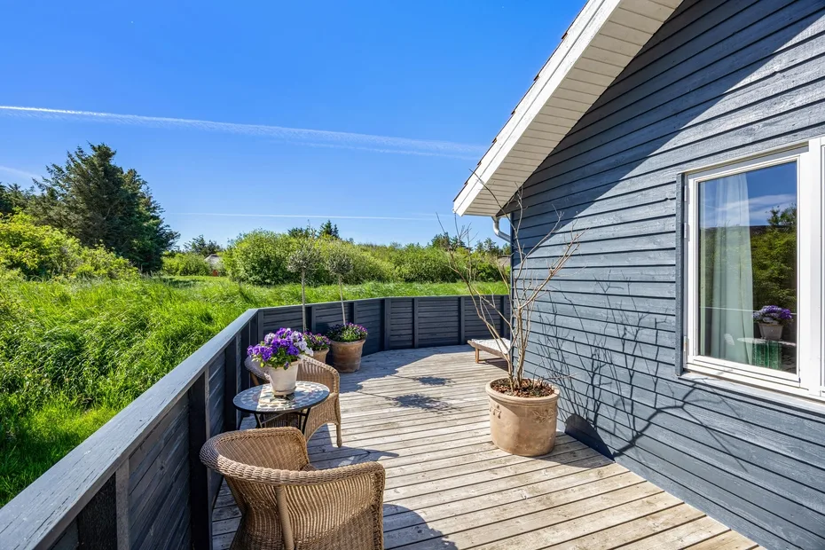 A sunny wooden deck beside a house, with outdoor seating and potted plants.