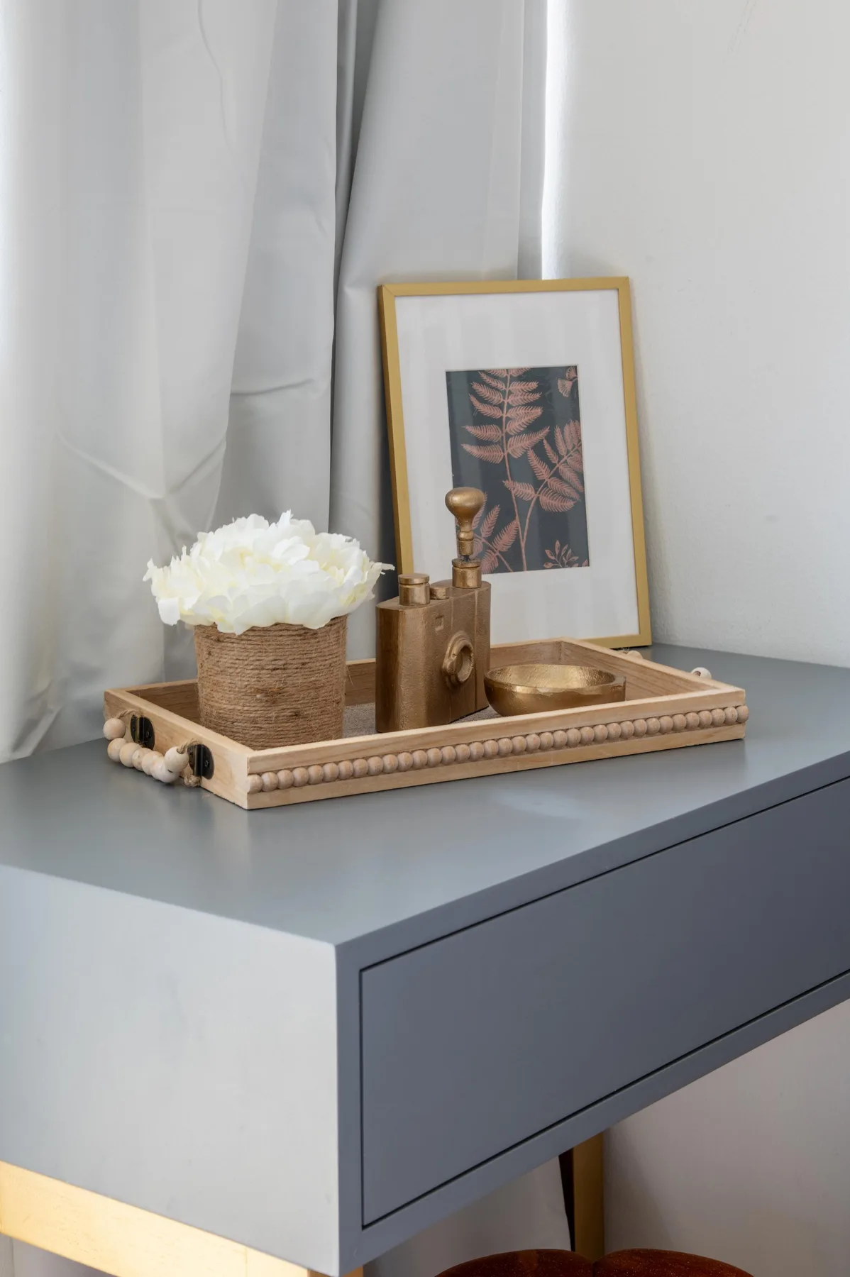 Un pequeño arreglo decorativo está sobre un mueble gris junto a una ventana con cortinas blancas.