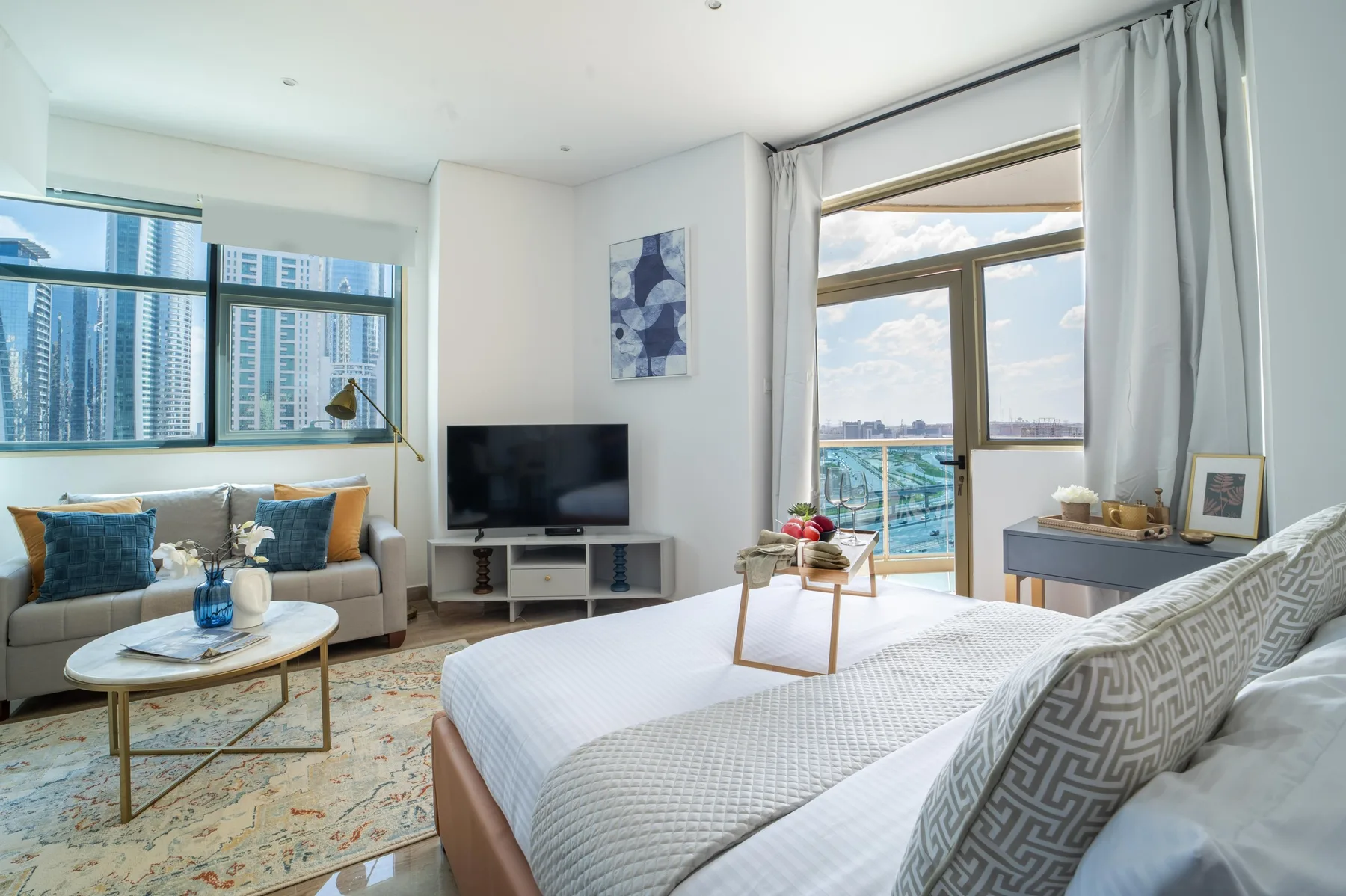 Un dormitorio luminoso con una cama grande, una zona de estar y ventanas con vistas a edificios cercanos.