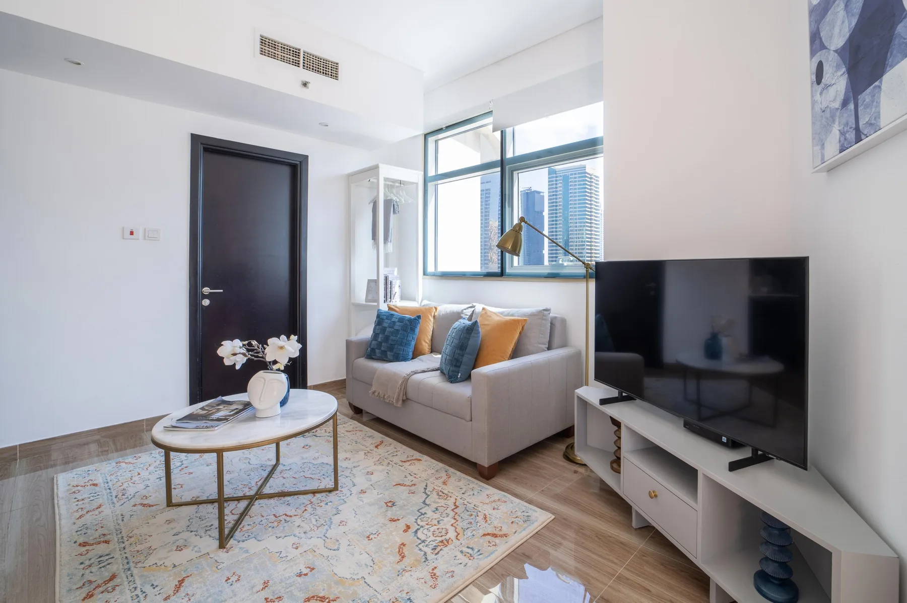 Una sala luminosa con sofá, mesa de centro y televisor junto a grandes ventanas.
