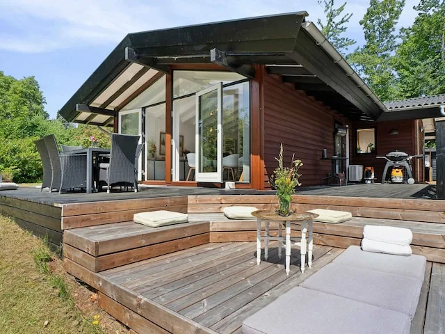A wooden house opens onto a raised deck with outdoor seating and a small table.