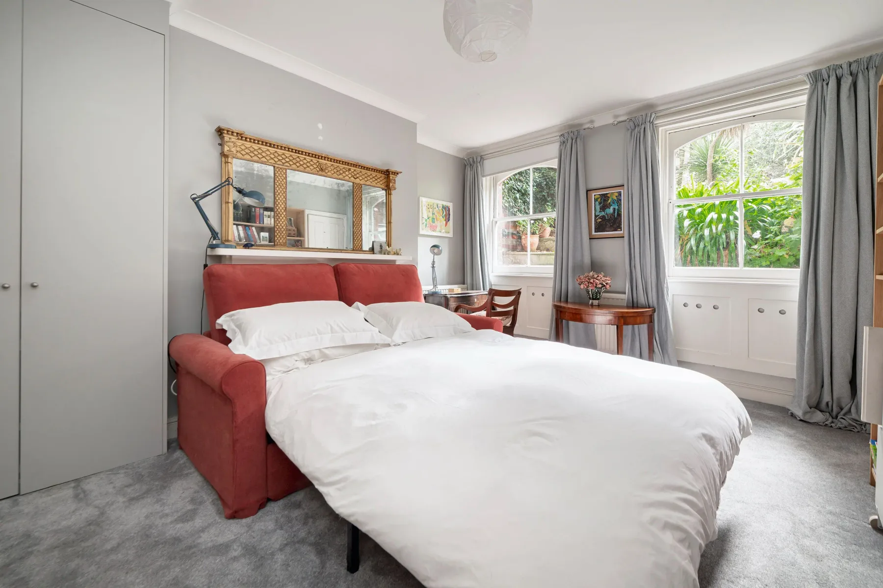 Un dormitorio luminoso con una cama tapizada roja y ventanas altas.