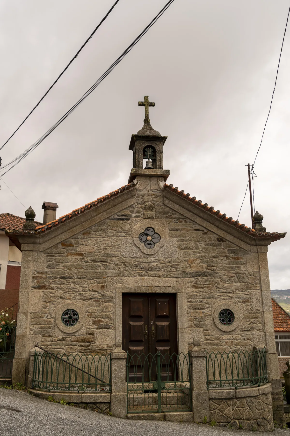 Una capilla de piedra con una torre de campanario coronada por una cruz está junto a una calle pavimentada.