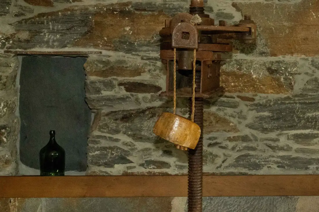Una pared interior de piedra rústica muestra una prensa de madera antigua y una cesta colgante.