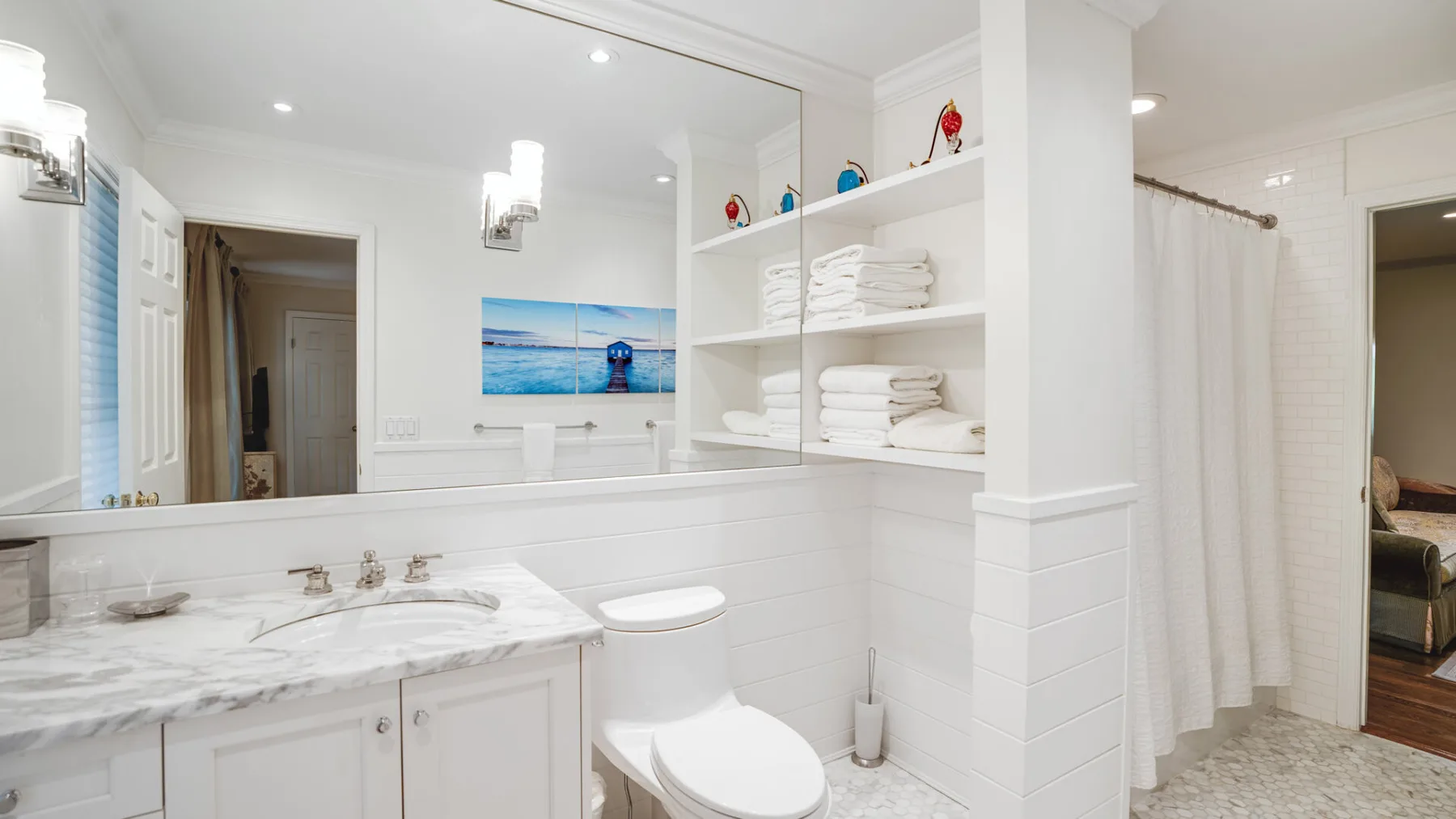 A bright bathroom with a marble vanity, toilet, and open shelves of folded towels.