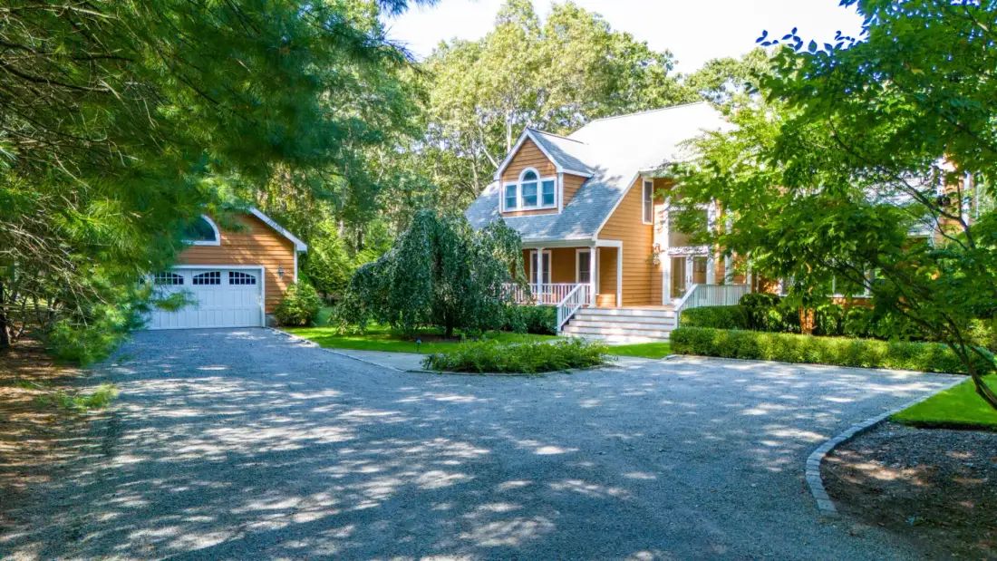 A house and detached garage sit beside a broad paved driveway, surrounded by trees.