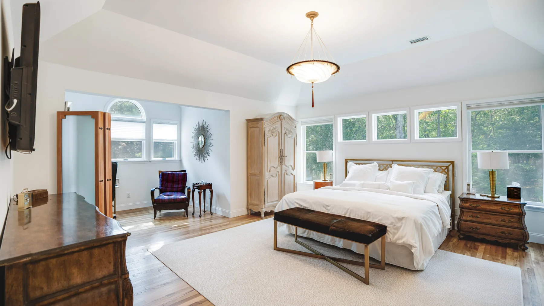 A bright bedroom with a large bed, a bench, and windows along the wall.