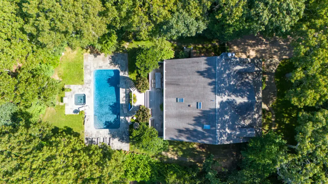 An aerial view shows a house beside a swimming pool, surrounded by trees.