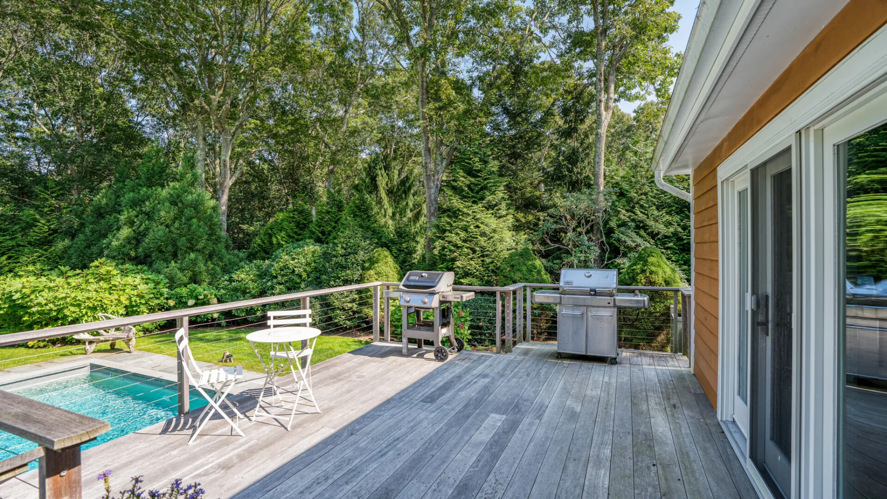 A wooden deck with outdoor seating and grills overlooks a swimming pool and leafy trees.