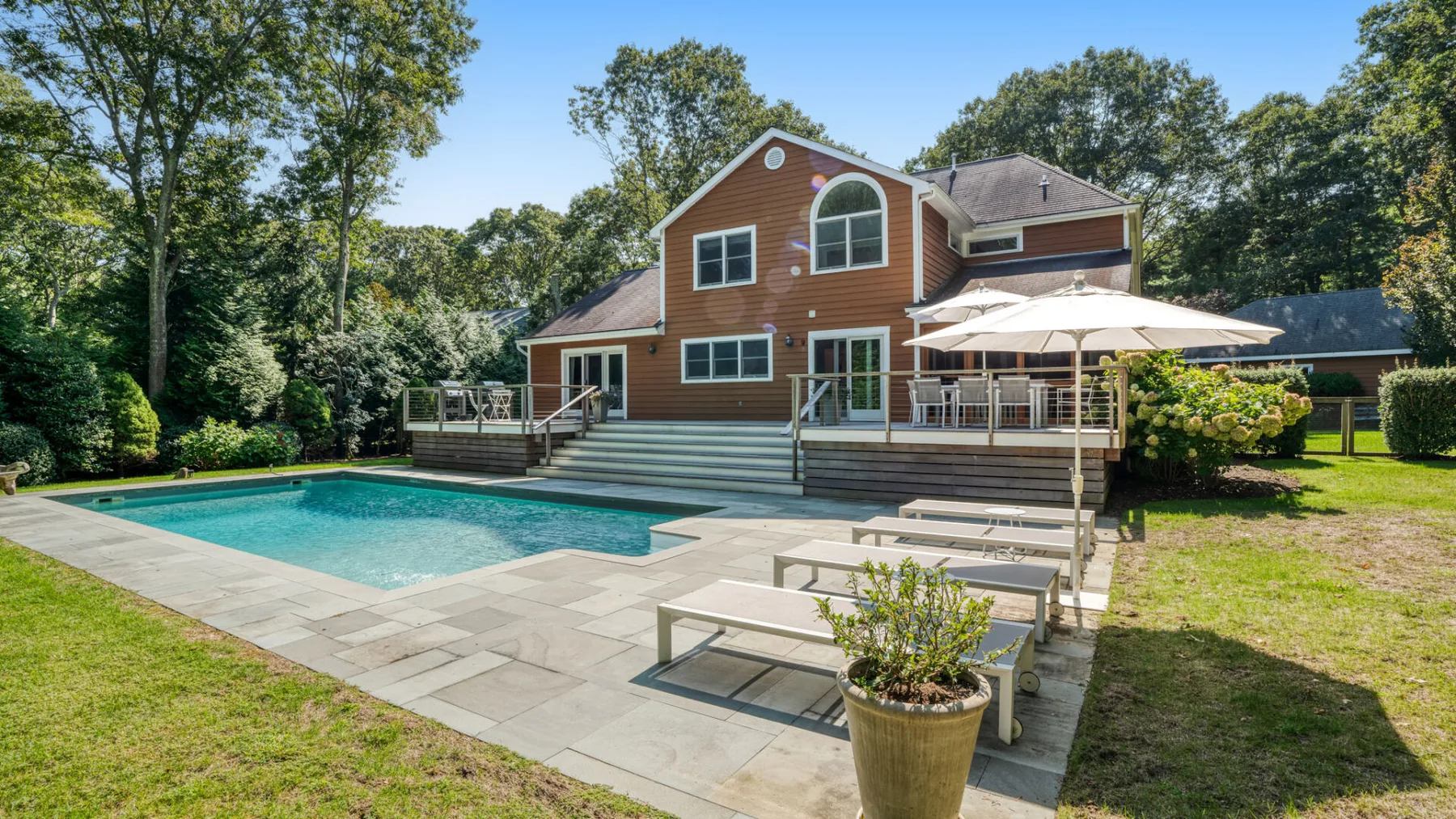 A backyard pool and patio sit beside a wood-sided house surrounded by trees.