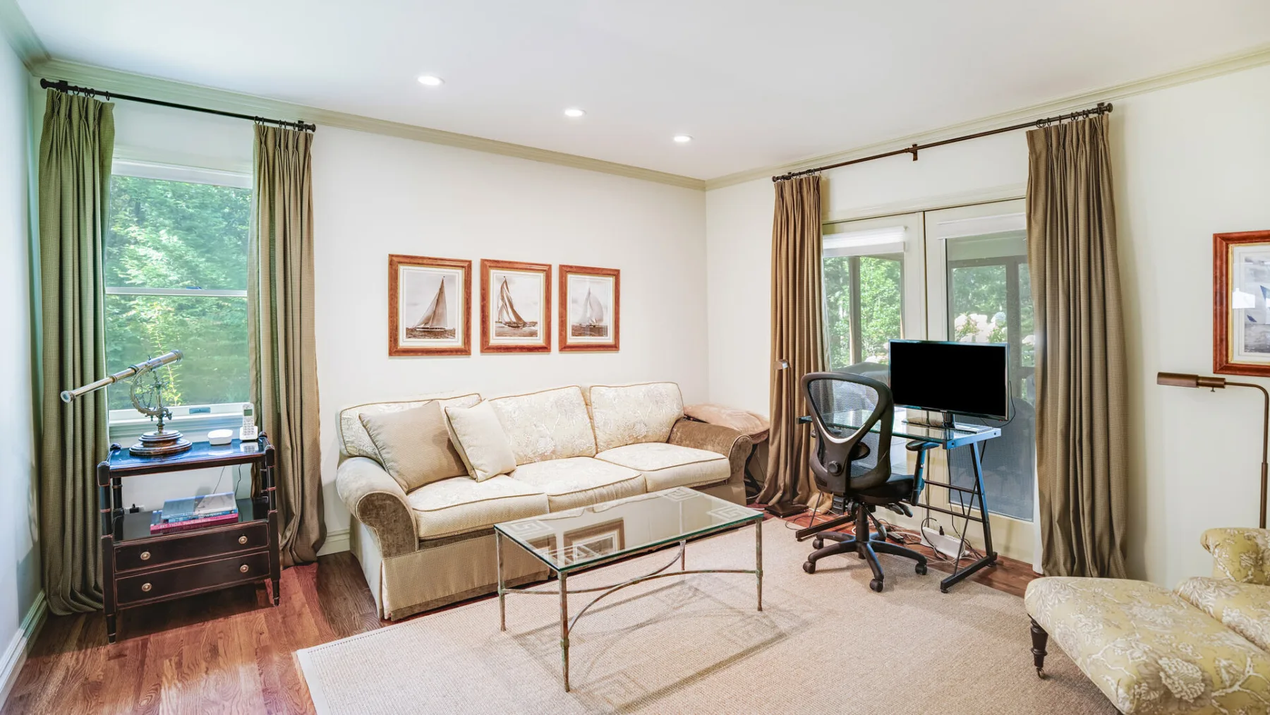 A bright living room with a cream sofa, glass coffee table, and desk by the windows.