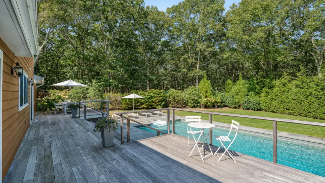 Una piscina junto a una terraza de madera, con asientos al aire libre y árboles alrededor.