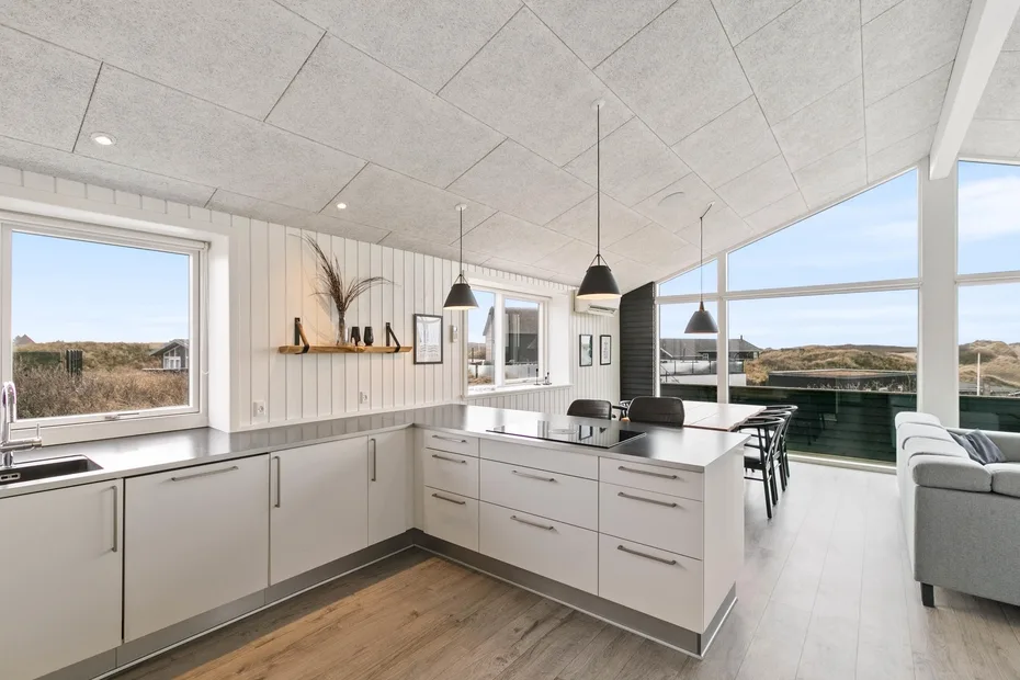 Una cocina abierta y luminosa con gabinetes blancos, lámparas colgantes y grandes ventanas.