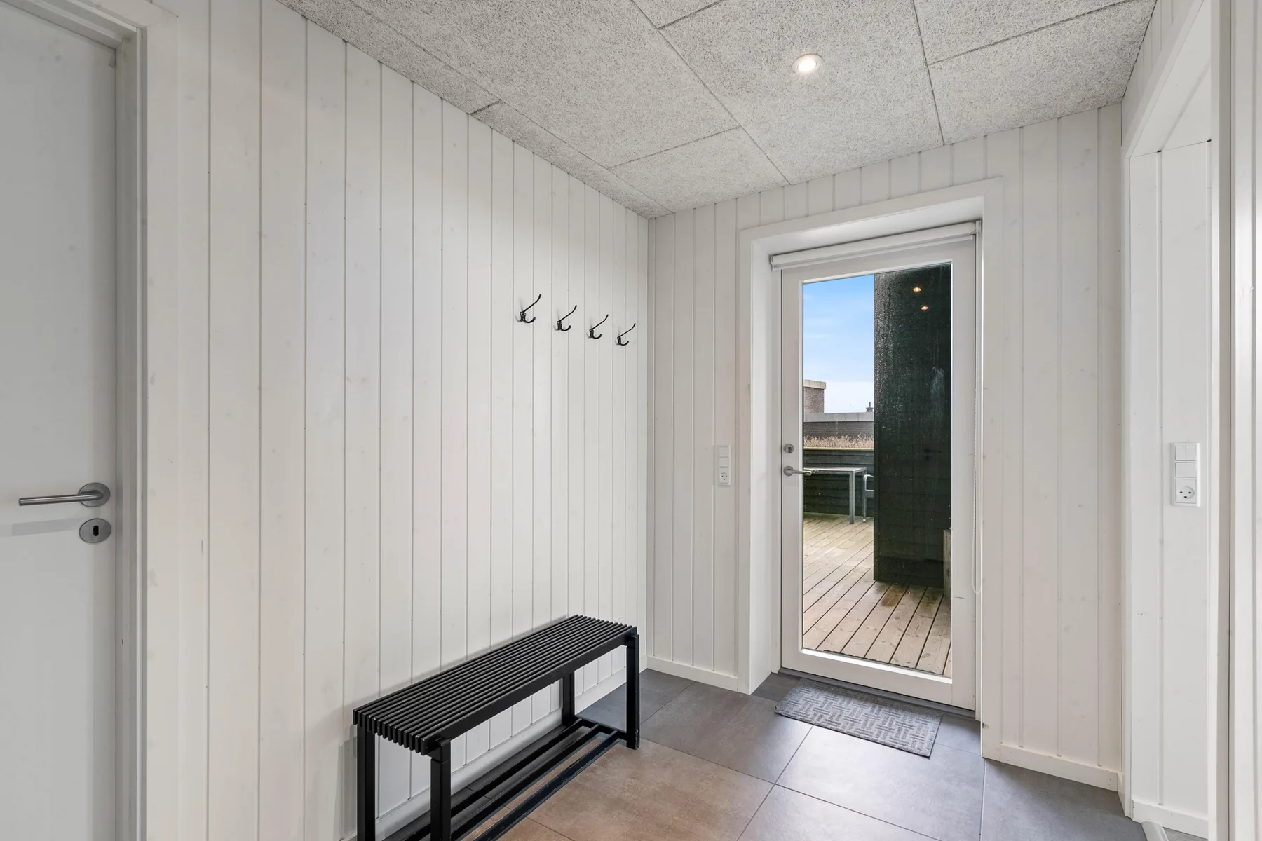 Una entrada luminosa con un banco, ganchos de pared y una puerta de vidrio hacia una terraza.