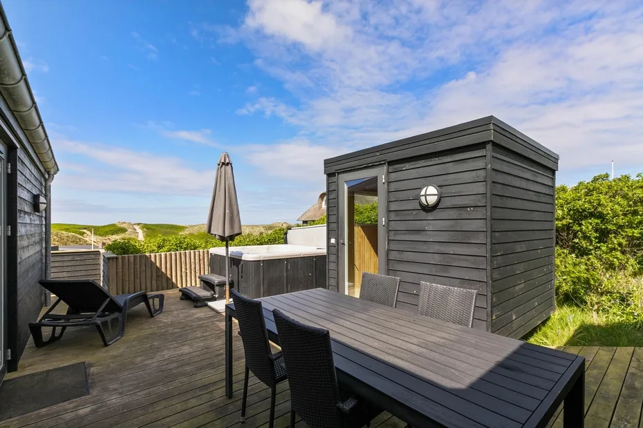 Una terraza de madera tiene asientos al aire libre, una mesa y una bañera de hidromasaje junto a un cobertizo.