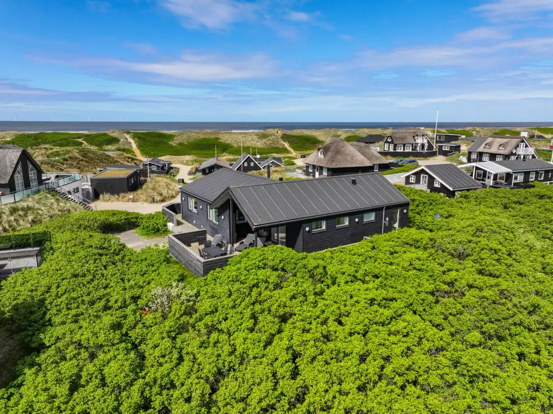 Una casa moderna oscura está entre dunas verdes, con el mar visible a lo lejos.