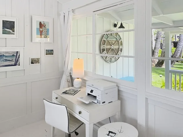 Un escritorio blanco y luminoso está junto a grandes ventanas, con cuadros en la pared.