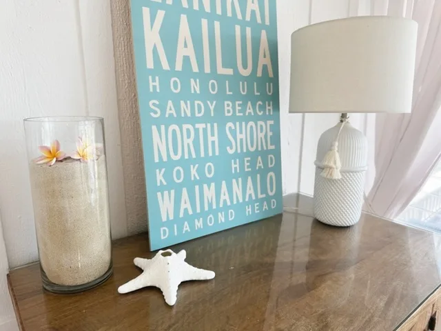 Una mesa consola de madera exhibe un cuadro con nombres de playas, una lámpara y una estrella de mar.