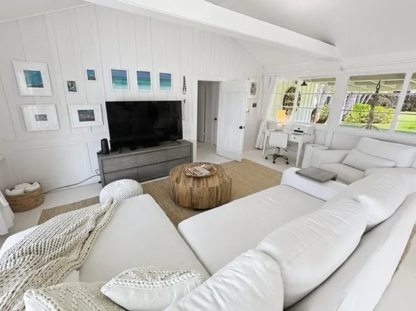 Una sala luminosa con asientos blancos, un televisor y ventanas grandes.