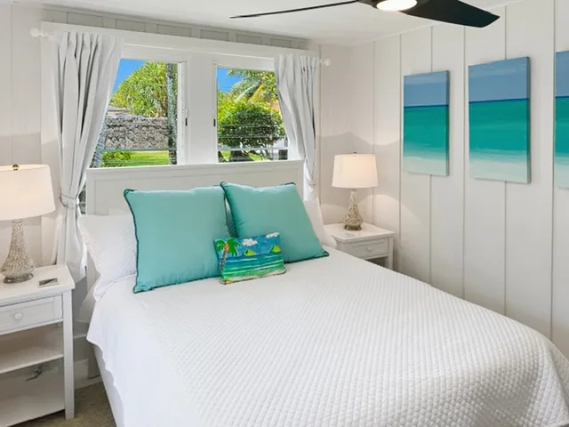 Un dormitorio luminoso con una cama blanca, cojines turquesa y una ventana con vistas a la vegetación.