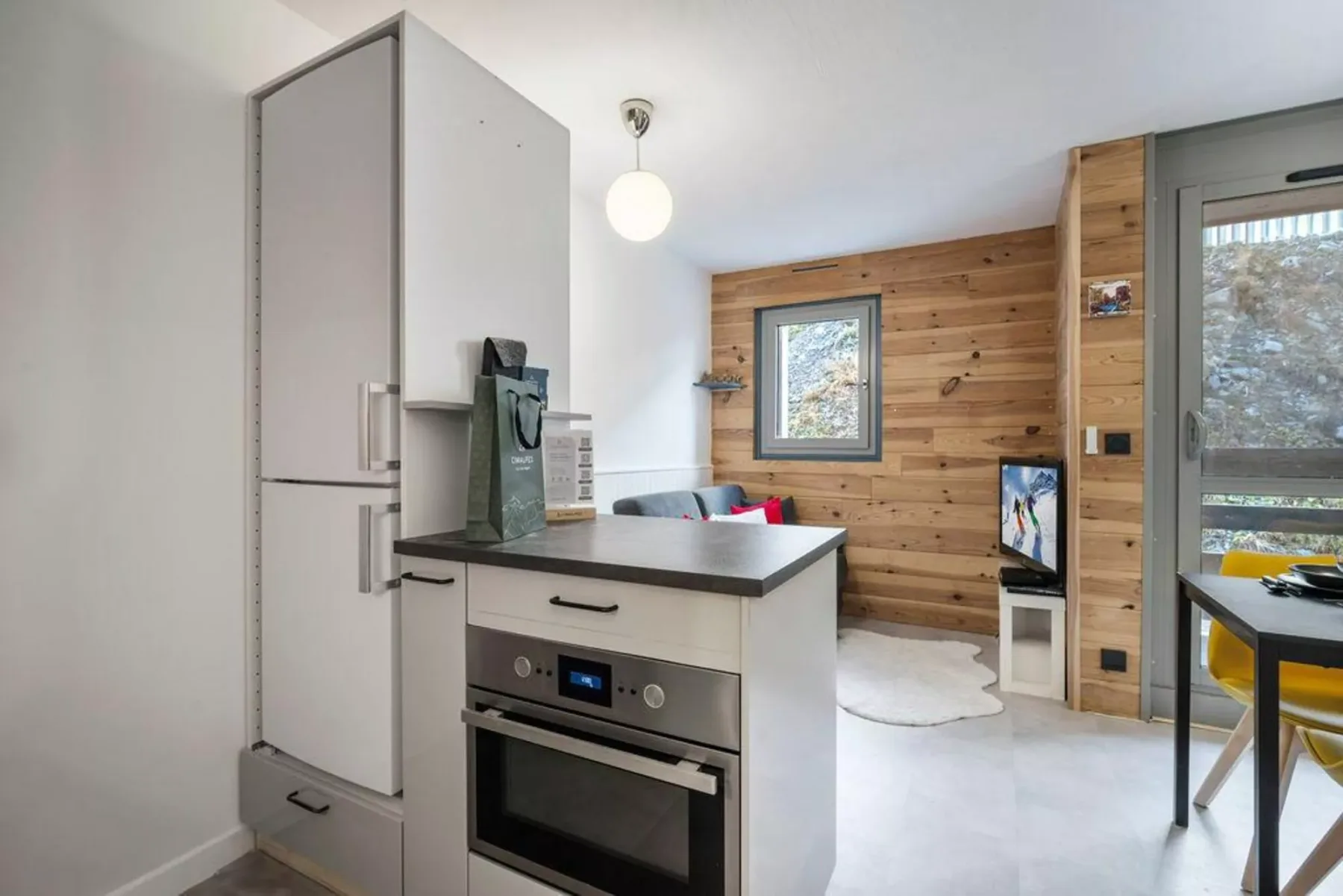 Una cocina compacta se abre a una sala con paredes de madera, ventana y televisor.