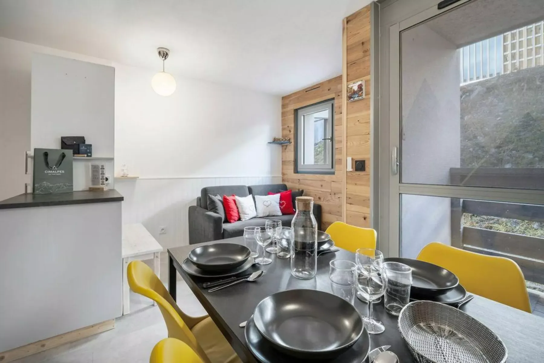 Comedor con la mesa puesta junto a una pequeña zona de estar y ventanas.