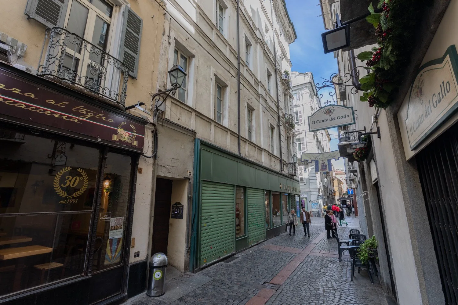 A narrow cobblestone street runs between tall buildings with storefronts and café signs.