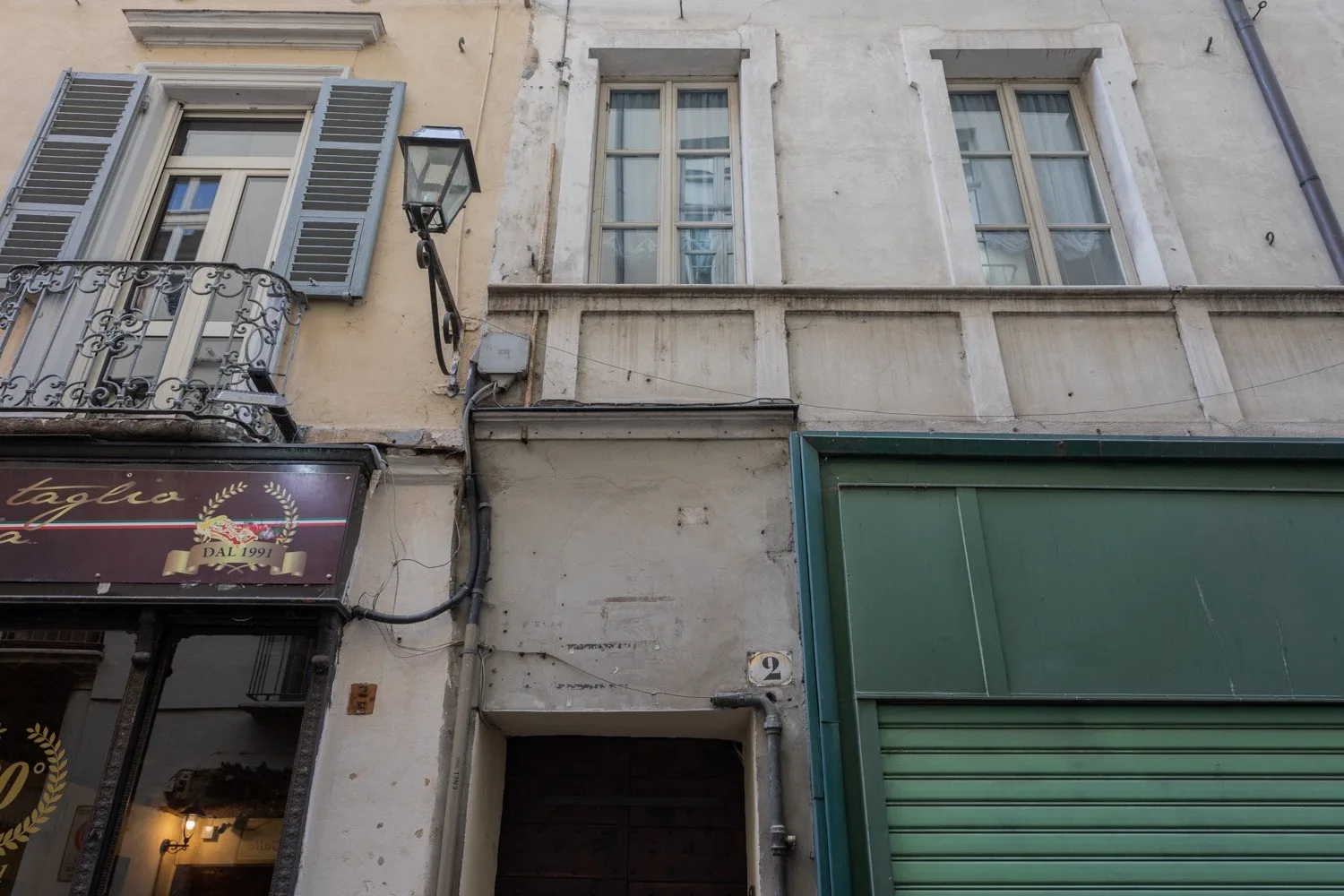 A narrow street-facing building facade has upper windows and a green rolling shutter.