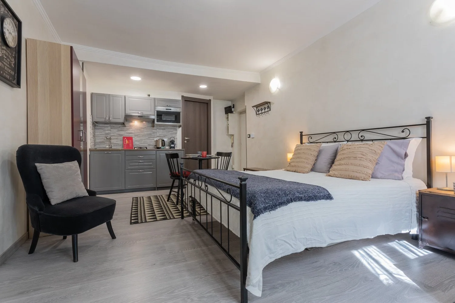 A bedroom with a bed, seating area, and compact kitchen in the background.
