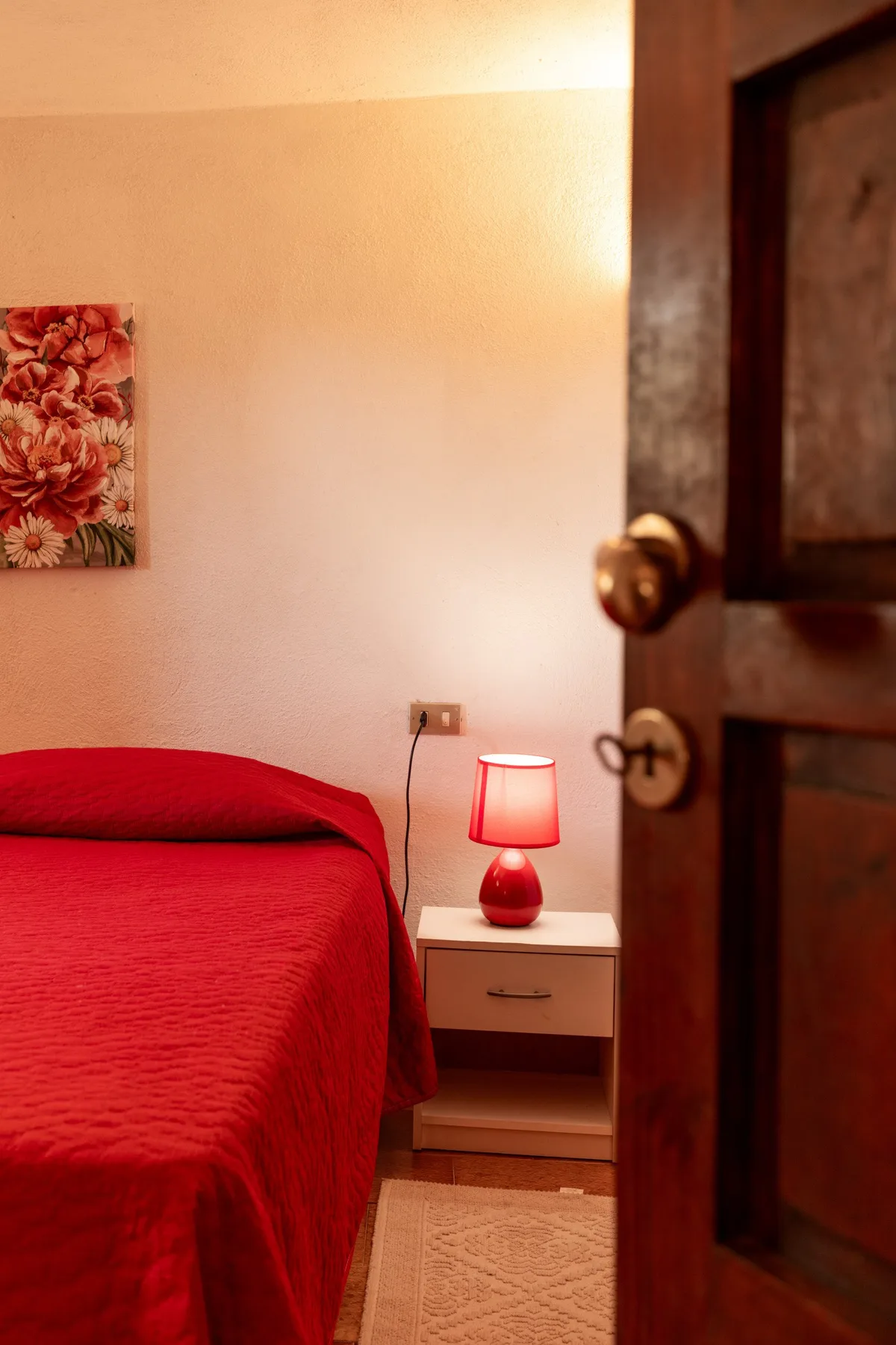 A bedroom with red bedding, a bedside table, and a lit lamp.